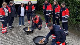 Mehrere Personen in Malteser Kleidung stehen um zwei Feuerschalen, an denen zwei Personen ein Feuer entzünden
