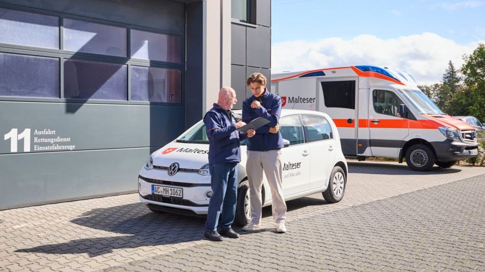 Zwei Menschen stehen vor einem Fahrzeug der Malteser und unterhalten sich