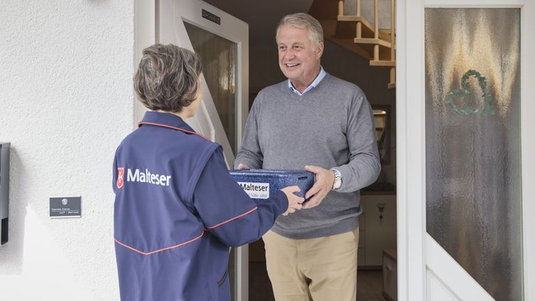 Eine Mitarbeiterin des Malteser Menüservice übergibt eine Mahlzeit an der Tür. 
