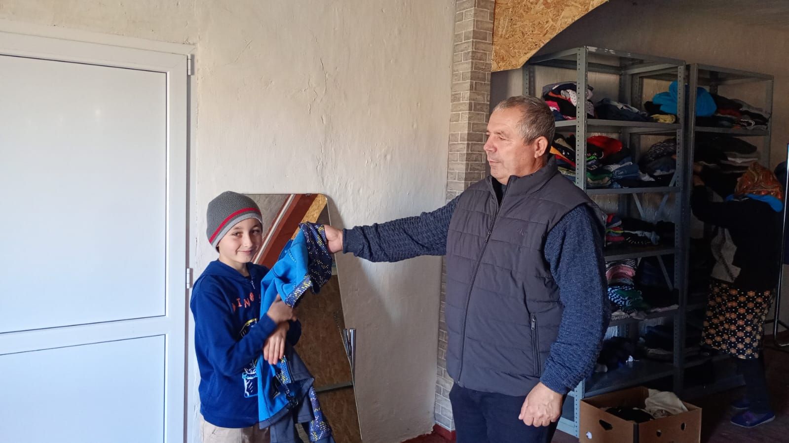 Ein Junge steht vor einer weißen Wand und erhält von einem älteren Mann eine neue Jacke aus der Kleiderkammer