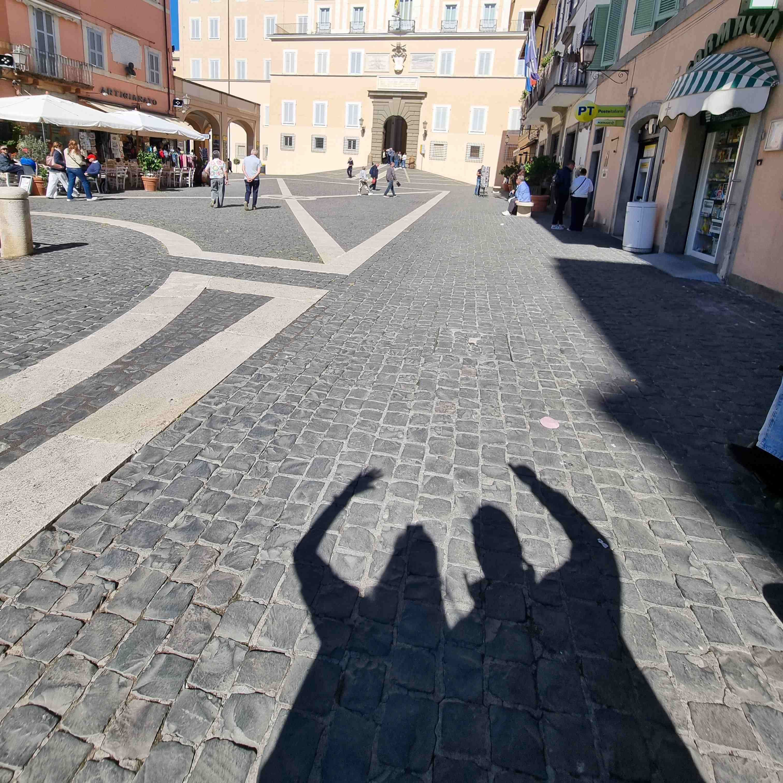 Schatten zweier Personen auf gepflastertem Platz mit weißen Linien, umgeben von Gebäuden in Castel Gandolfo.