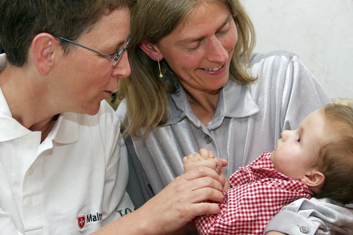 Mitarbeiterin der Malteser lacht Kind an, das im Arm der Mutter liegt.