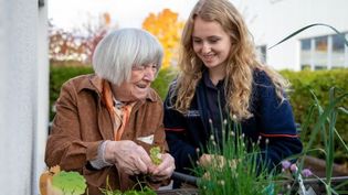 Eine Juniordemenzbegleiterin steht mit einer älteren Dame auf dem Balkon im Pflegeheim. Beide unterhalten sich. Die Demenzbegleiterin lächelt.