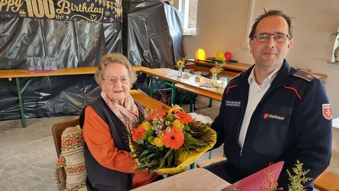 Die Jubilarin sitzt mit einem Blumenstrauß in der Hand an einem Tisch, daneben sitzt Malteser Kreisgeschäftsführer Roland Bucher