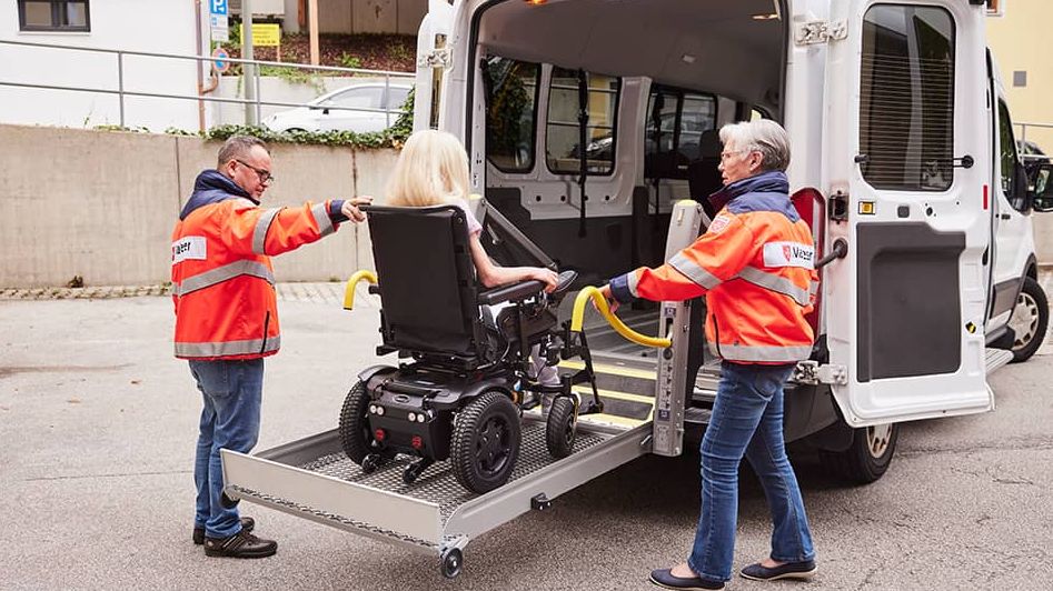 Zwei Malteser helfen einem Mädchen im Rollstuhl in ein Fahrzeug.