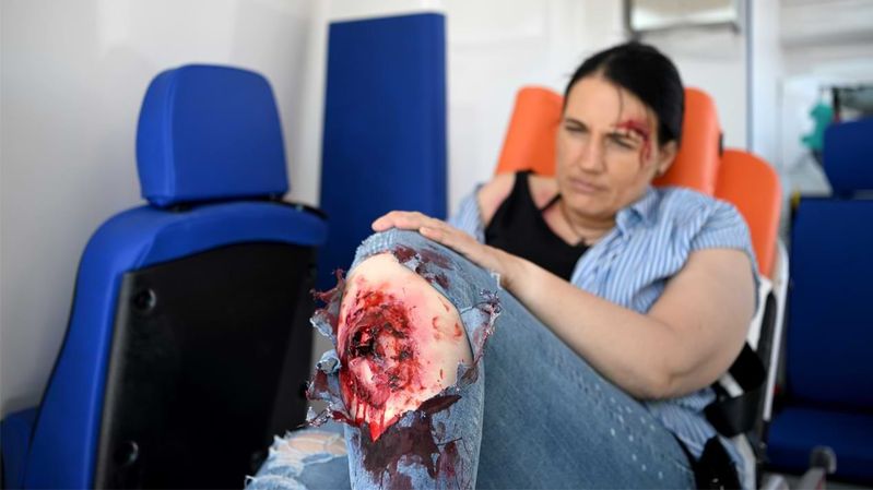 Eine verletzte Frau, mit täuschend echten Wunden an Stirn und Bein, liegt in einem Krankenwagen.