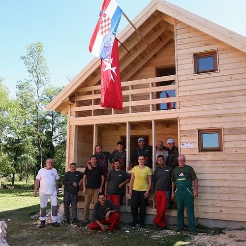 Eine Gruppe von ehrenamtlichen Helfern steht vor einem Holzhaus mit Malteser und Kroatien Flagge. 