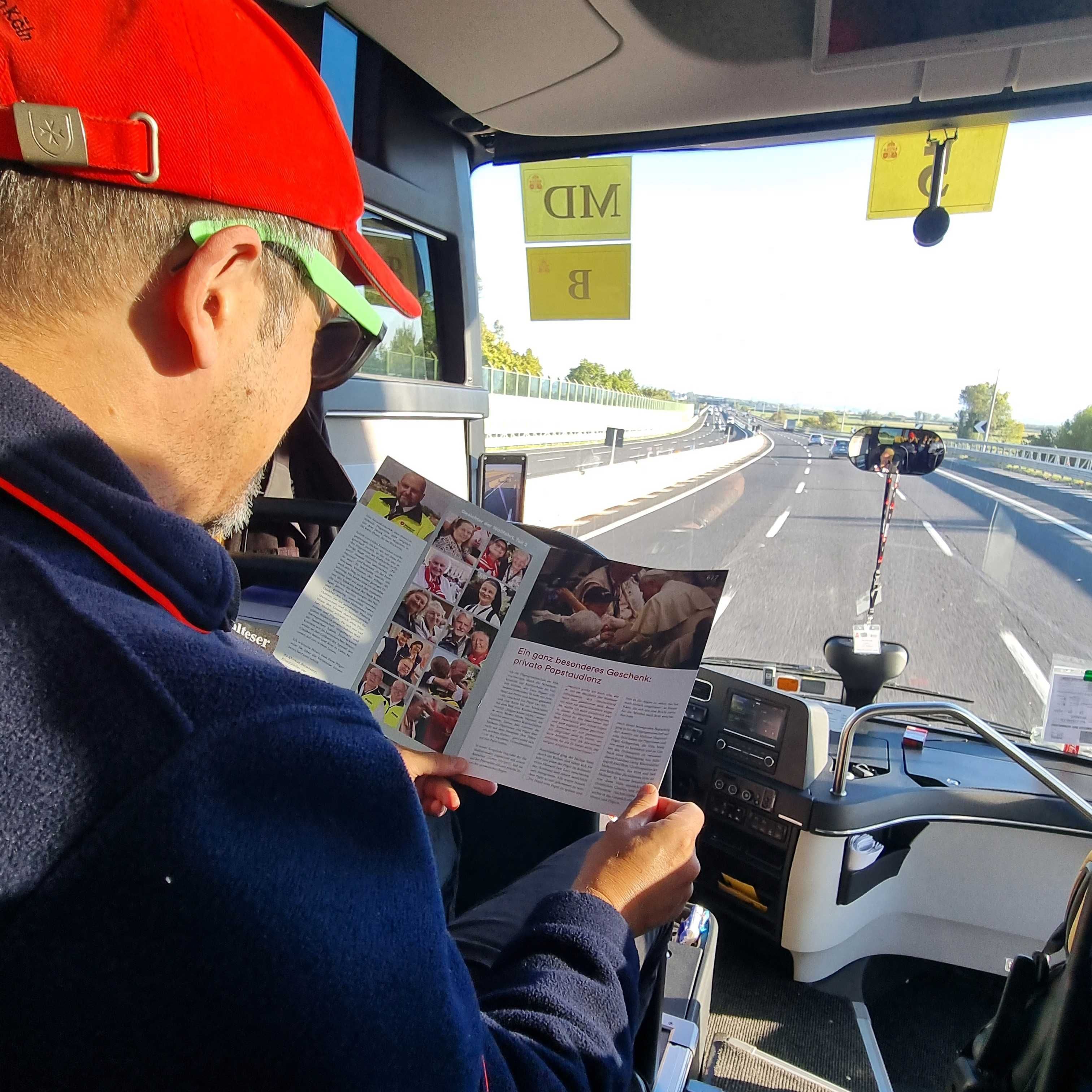 Person mit roter Kappe und dunkler Jacke liest Broschüre im Reisebus eines Fahrzeugs, Blick auf Straße mit mehreren Fahrzeugen