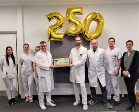 Team in weißer Dienstkleidung feiert gemeinsam 250. OP mit Kuchen und goldenen Luftballons in Form der Zahl 250.