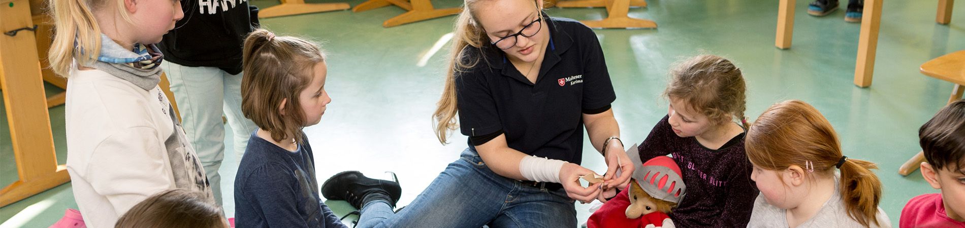 Das Bild zeigt eine Szene in einem Raum der offenen Ganztagsbetreuung. Eine Erzieherin sitzt auf dem Fußboden und umringt von Kindern. Sie "verarztet" ein Plüschtier mit Verbandsmaterial das von einem Mädchen gehalten wird. Die Kinder scheinen neugierig zu sein und beobachten die Erzieherin aufmerksam. Der Raum ist mit Spielzeug und Möbeln für Kinder eingerichtet.