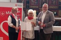 Eine Frau zeigt eine Urkunde, neben ihr stehen ein Mann und eine Frau in Malteser Kleidung.