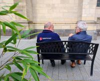 Ein Malteser-Ehrenamtler sitzt auf einer Bank im Freien mit einem älteren Herrn. Sie unterhalten sich.