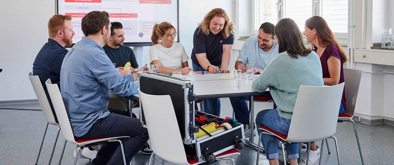 Eine Ausbilderin sitzt mit Teilnehmenden der Erste-Hilfe-Fortbildung an einem Tisch.