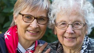 Malteser Mitarbeiterin legt Arm um ältere Frau und beide lachen in die Kamera.