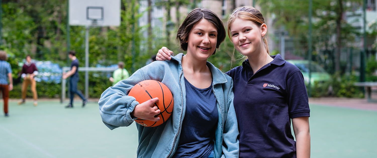 Eine junge Frau im Malteser Shirt legt einem Mädchen mit einem Basketball den Arm um die Schulter.