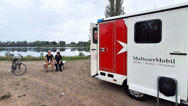 Das MalteserMobil ist zu sehen, ein Auto für die Obdachlosenhilfe. Zwei Frauen sitzen daneben auf einer Bank. Im Hintergrund ist Wasser zu sehen.