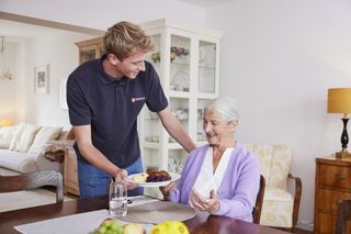 Ältere Frau in lilafarbenem Oberteil an einem Esstisch, junger Mann in Malteser Polohemd reicht ihr Essen auf einem Porzellanteller, 