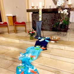 Ein blaues Tuch liegt vor dem Altar in einer Kirche. Das Tuch symbolisiert einen Fluss. Auf dem Fluss stehen kleine Boote aus Papier. Darauf sind Namen geschrieben.