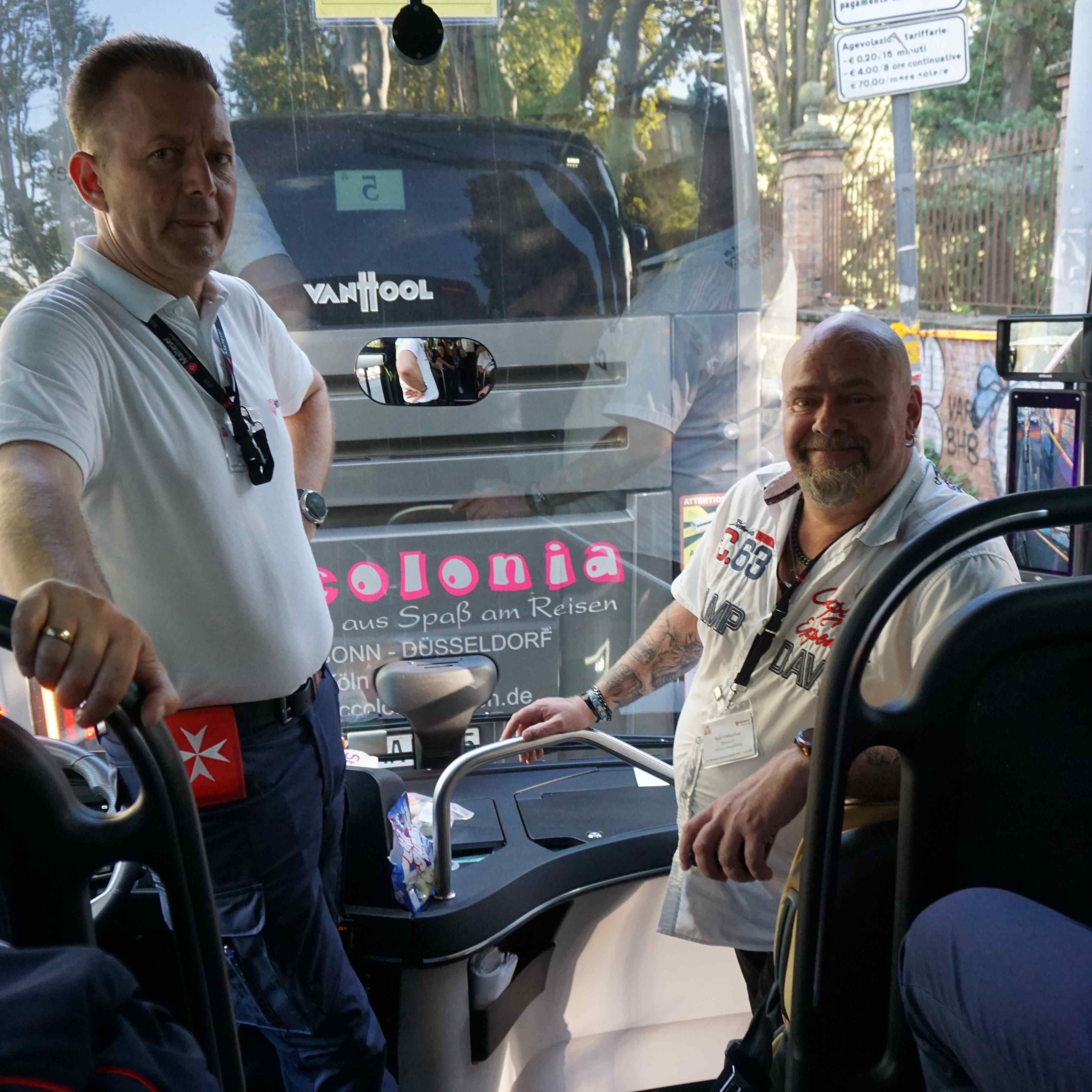 Zwei Männer stehen und sitzen im vorderen Bereich eines Busses, im Hintergrund ist die Front eines Busses mit Schriftzug sichtbar.