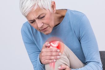 Seniorin hält sich mit schmerzverzerrtem Gesicht das Knie, rotes Symbol markiert Schmerzstelle.
