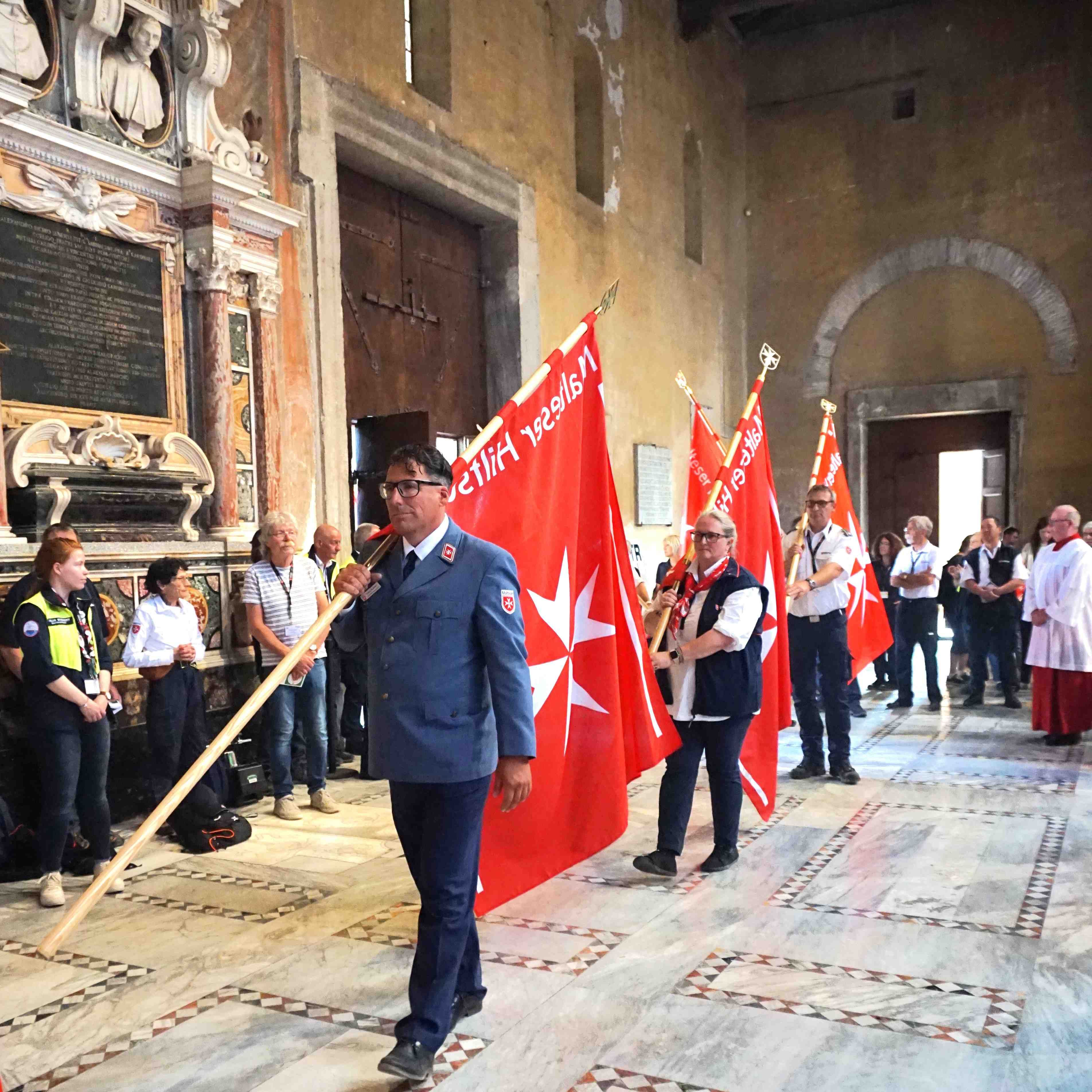 Im Mittelpunkt läuft eine männliche Person in einer Reihe und trägt eine Malteser Fahne am Banner.