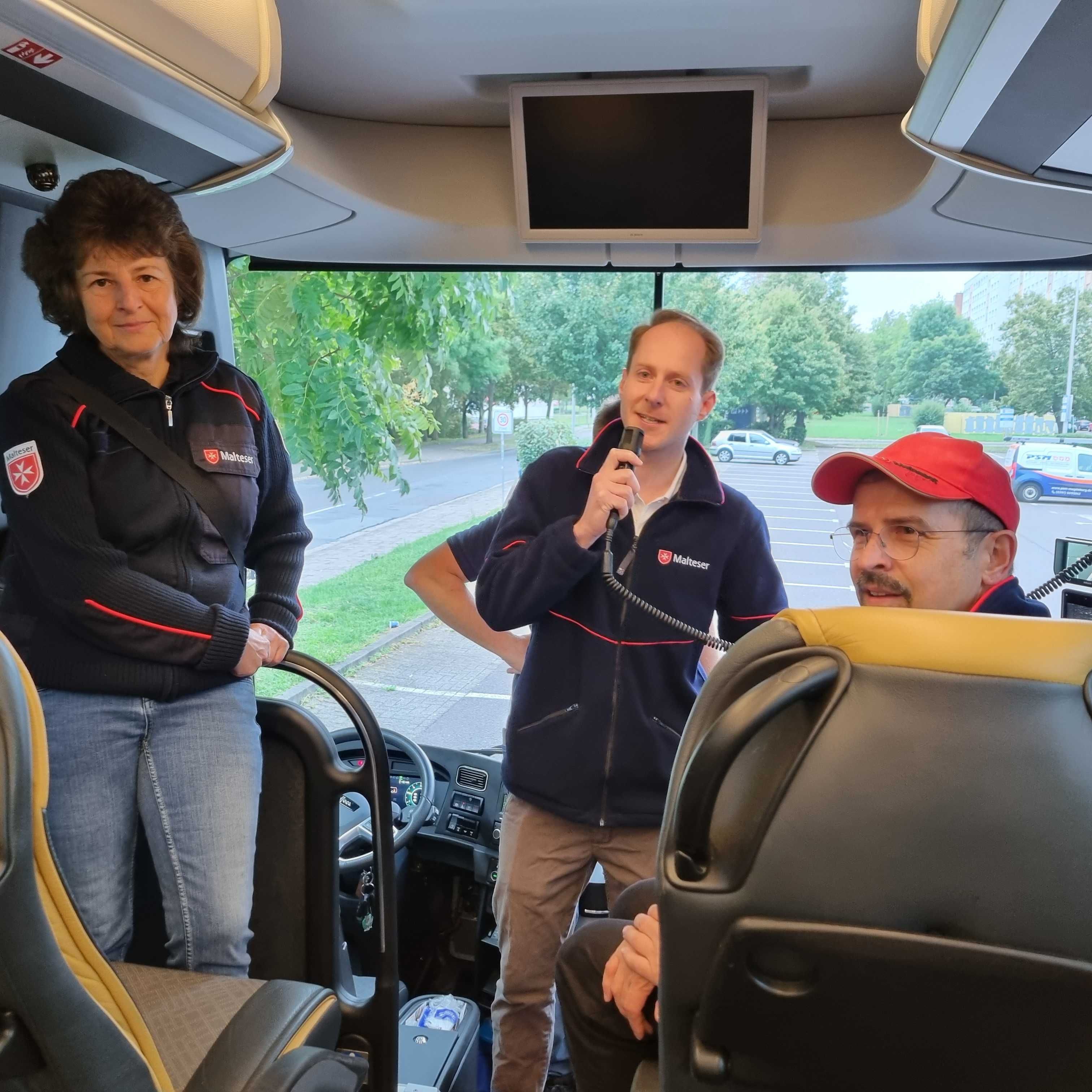 Person mit dunkelblauer Malteser Jacke und Mikrofon steht im Bus vorne neben Fahrersitz, weitere Personen sitzen und stehen im Bus.