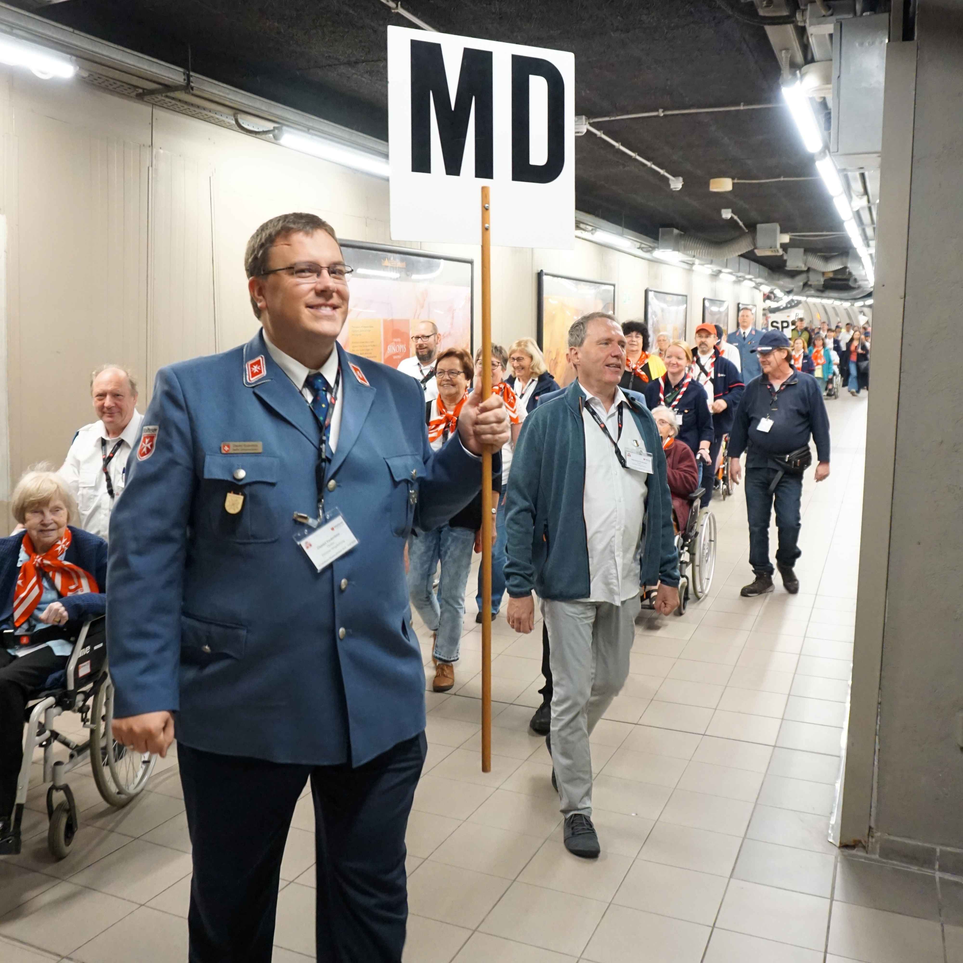 Person in Malteser Uniform führt Gruppe von Menschen durch einen Gang, eine Person hält ein Schild mit der Aufschrift 'MD'.