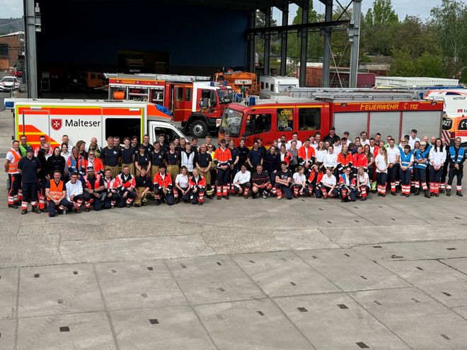 Eine große Gruppe von Einsatzkräften aus Feuerwehr und Rettungsdienst steht gemeinsam vor mehreren Einsatzfahrzeugen auf einem Gelände. Im Hintergrund sind ein Feuerwehrfahrzeug und ein Rettungswagen der Malteser zu sehen.