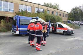 Auf einem Parkplatz gehen mehrere Einsatzkräfte in Schutzkleidung zu einem blauen Mannschaftsfahrzeug und einem weißen Einsatzfahrzeug. Im Hintergrund steht ein mehrstöckiges Gebäude, weitere Fahrzeuge sind am Fahrbahnrand geparkt.