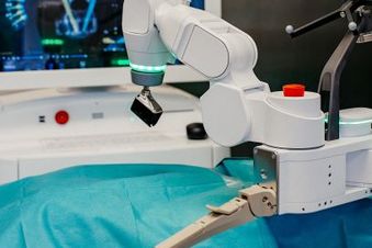 OP-Roboterarm steht über einem abgedeckten Operationstisch, im Hintergrund ist ein Monitor zu sehen.