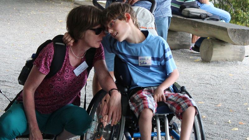 Eine hockende Frau mit Sonnenbrille hält ihren Kopf an den Kopf eines Jungen, der in einem Rollstuhl sitzt.