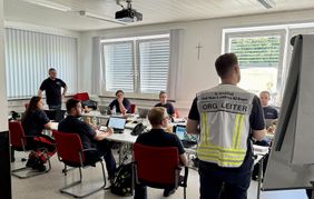 Ausbildungsraum, in dem die Übung stattfindet. Die Teilnehmenden sitzen an Tischen, der Übungsleiter steht mit dem Rücken zur Kamera.
