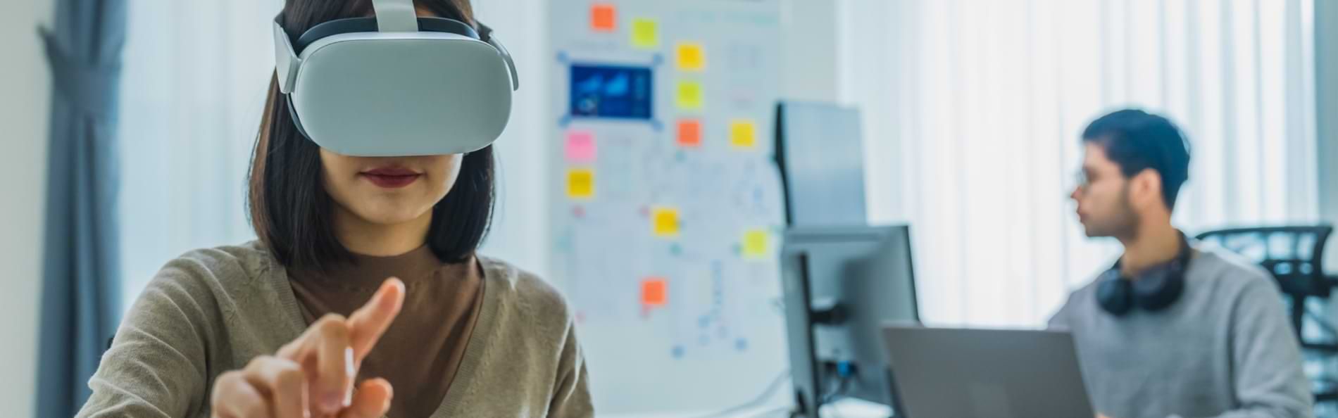 Eine Frau mit VR-Brille im Vordergrund, dahinter ein Mann mit Laptop und einem Whiteboard.