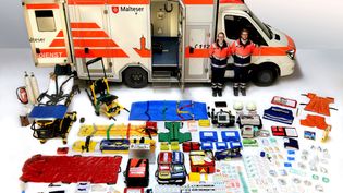 Rettungswagen der Malteser mit offener Tür, zwei Rettungskräfte in Uniform, zahlreiche medizinische Geräte und Ausrüstungsgegenstände ordentlich vor dem Fahrzeug ausgelegt.