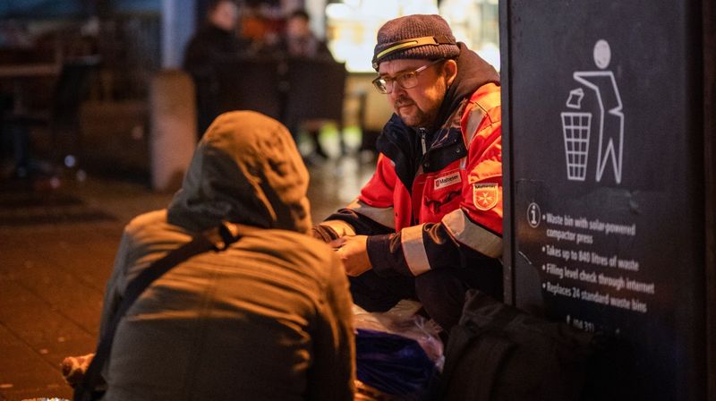 Ein Mann in Einsatzkleidung der Malteser kniet vor einer Person, die auf dem Bürgersteig sitzt.