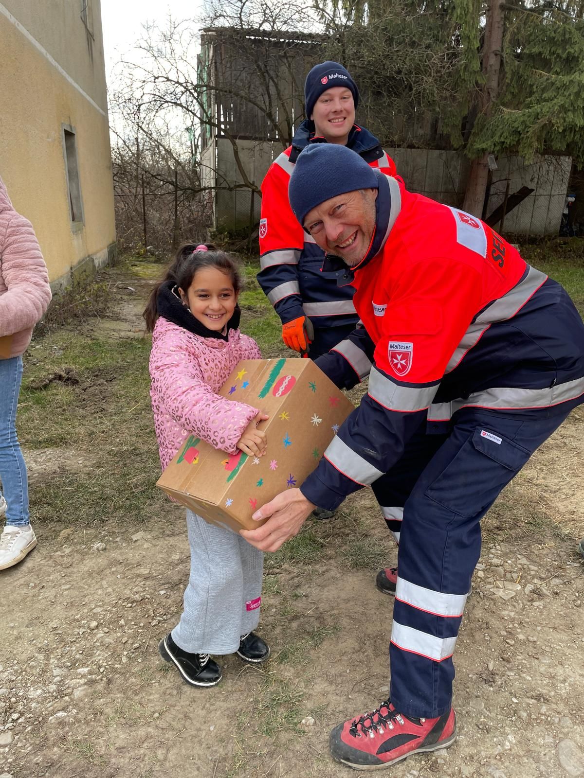 Mann in Malteser Kleidung überreicht einem kleinen Mädchen ein großes Paket
