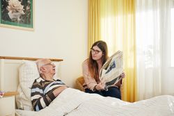 Eine junge Frau sitzt an einem Bett, in dem eine ältere Person liegt.