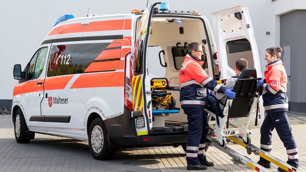 Malteser in Einsatzkleidung helfen einem gehbehindertem Mann in in ein Fahrzeug.