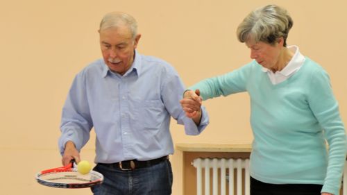 Älterer Mann balanciert Ball auf Tennisschläger, unterstützt von einer Begleitperson.