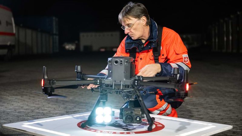 Ein Mann mit Brille und Einsatzkleidung der Malteser kniet vor einer Drohne und überprüft die Technik.