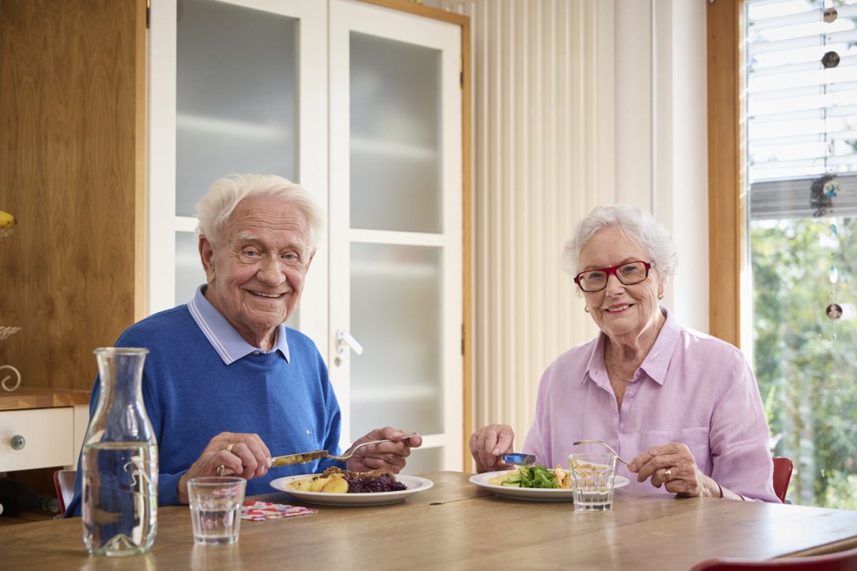 Älterer Mann im blauen Pulli und Seniorin in einem fliederfarbenen Oberteil sitzen vor Tellern mit warmem Essen