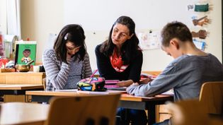 Drei Personen sitzen an einem Tisch in einem Klassenraum, umgeben von Schulmaterialien und Kinderzeichnungen an der Wand.