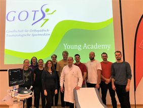 Gruppe mit Arzt vor Projektionsfläche mit GOTS-Logo und Text „Young Academy“ in einem Seminarraum.