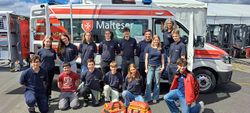 14 Schulsanis stehen oder knien gemeinsam vor einem Malteser Rettungswagen und schauen lächelnd in die Kamera. 