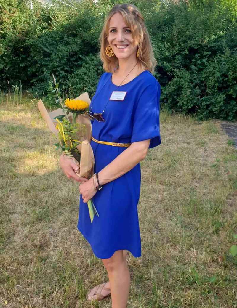 Eine lächelnde Frau in einem blauen Kleid mit einer Sonnenblume im Arm.
