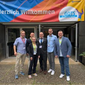 Sechs Teilnehmende stehen vor einem Kongressgebäude unter einem Willkommensbanner der GOTS.