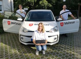 Drei Mitarbeitende des Malteser Hilfsdienst‑Menüservice stehen beziehungsweise knien vor einem weißen Dienstfahrzeug mit Malteser‑Logo. Links hält eine Frau ein Prospekt in der Hand, rechts steht ein Mann mit geöffnetem Fahrertür, vorne kniet eine Frau und hält eine isolierte Essensbox.