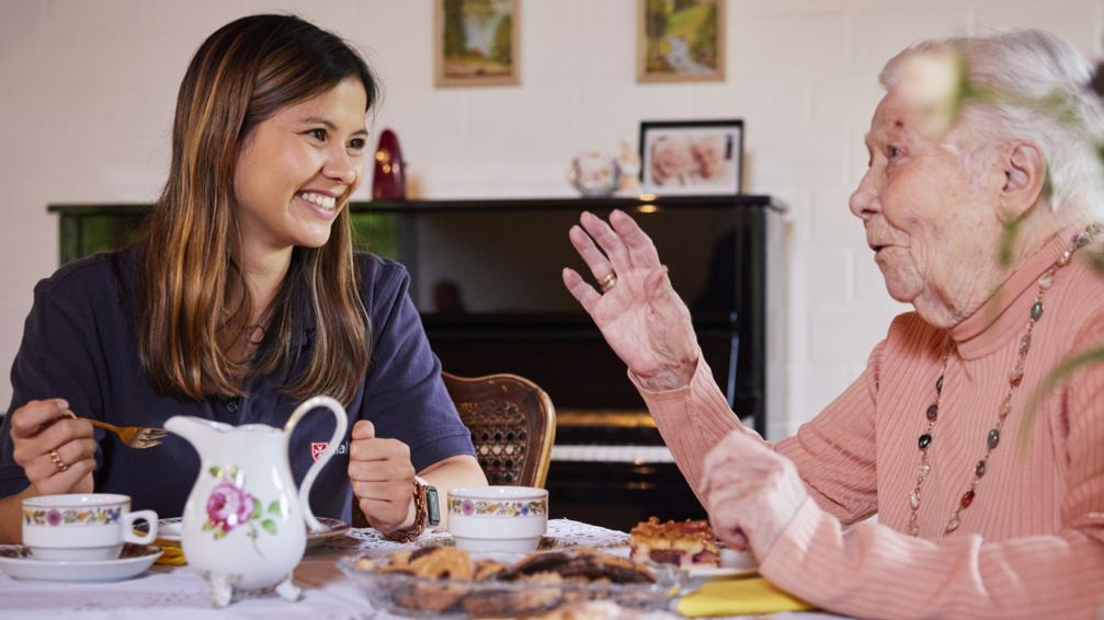 Junge Ehrenamtlerin des Besuchsdienstes sitzt mit einer Seniorin bei Kaffee und Kuchen zusammen.