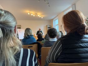 Mehrere Personen sitzen auf Stühlen in einem hellen Raum und hören einer vortragenden Person zu, die vorne an einem Tisch steht und aus einem Buch liest. Im Hintergrund befinden sich Regale mit Büchern und Ordnern sowie ein Flipchart mit einem Herz‑Motiv. Von der Decke hängen mehrere warm leuchtende Spotlampen, und durch die großen Fenster fällt Tageslicht in den Raum.