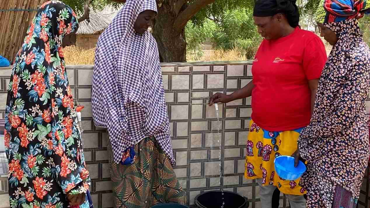 Eine Mitarbeiterin von Malteser International und drei weitere Frauen stehen im Freien um einen Wasserhahn und füllen Kunststoffeimer mit Wasser.